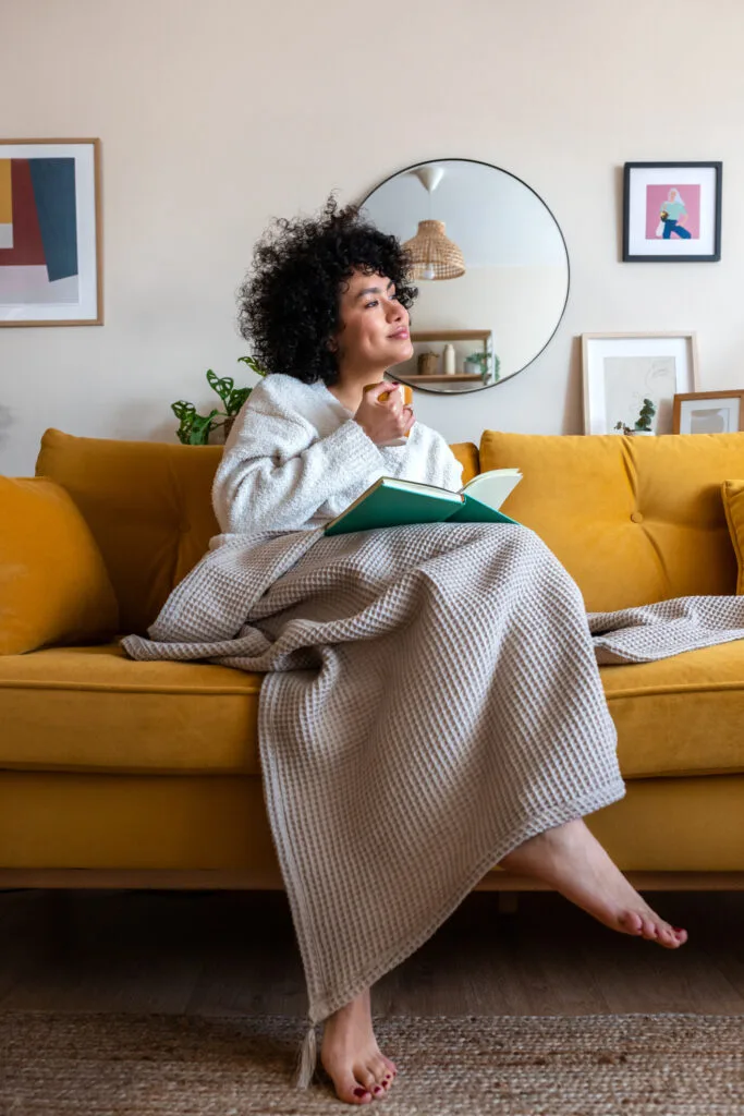 Pensive happy African american woman reading a book at home sitting on sofa relaxing drinking coffee. Vertical image