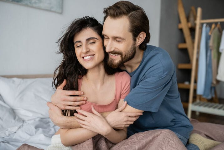 overjoyed-man-hugging-brunette-girlfriend-pajama-bed-home